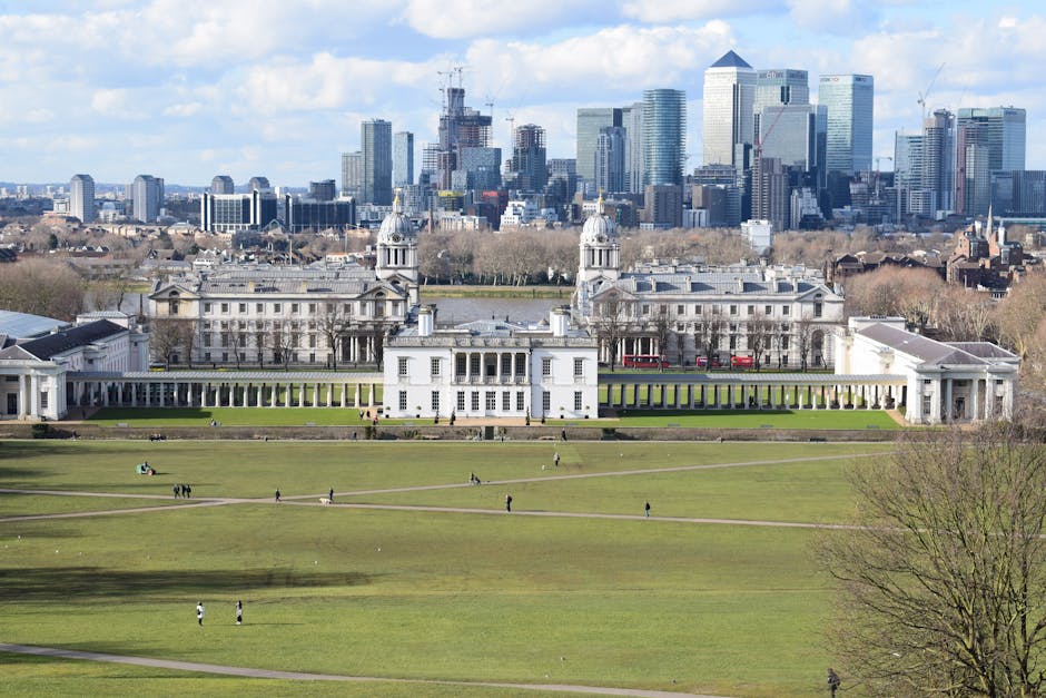 A wide view of the Royal Greenwich Park with well-maintained green lawns, pathways, and historic white buildings in the foreground, set against a modern London cityscape with tall office buildings and