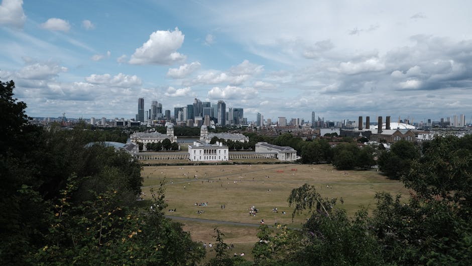 An expansive view of a historic white neoclassical building with ornate towers and colonnades, situated on a lush green park with walking paths and scattered trees in the foreground. Behind the buildi