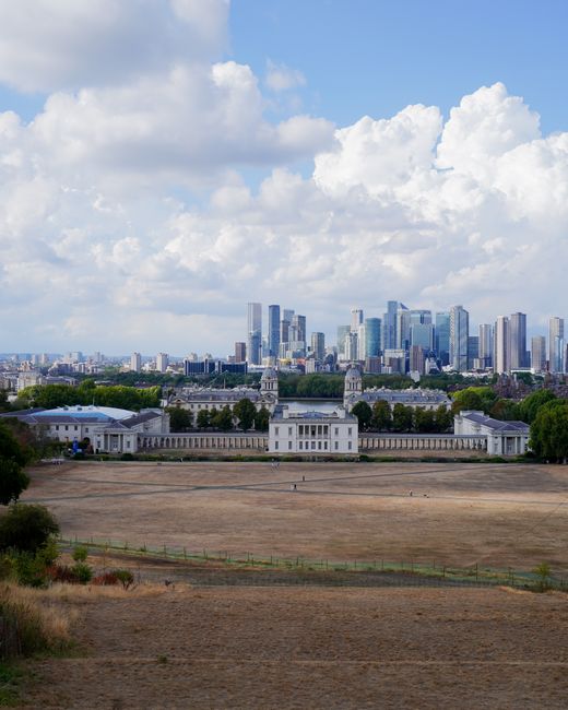 A landscape view of a wide, open field with dry, brown grass and a few small trees in the foreground. Beyond the field, there is a white historic building with classical architectural features, includ