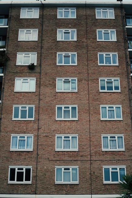 Photograph of a tall, multi-storey residential building constructed with brown brickwork, featuring numerous white-framed windows arranged in uniform rows. The building's façade appears clean and wel