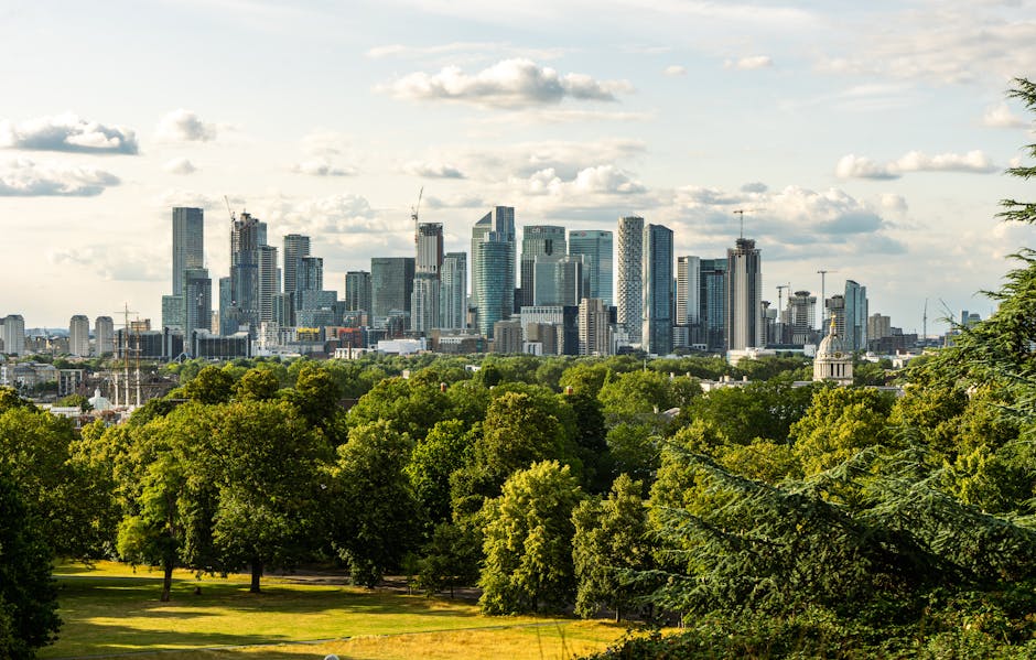 An expansive view of Greenwich Park with lush green lawns and scattered visitors sitting or walking. In the foreground, there is a white historic building surrounded by well-maintained gardens and pat