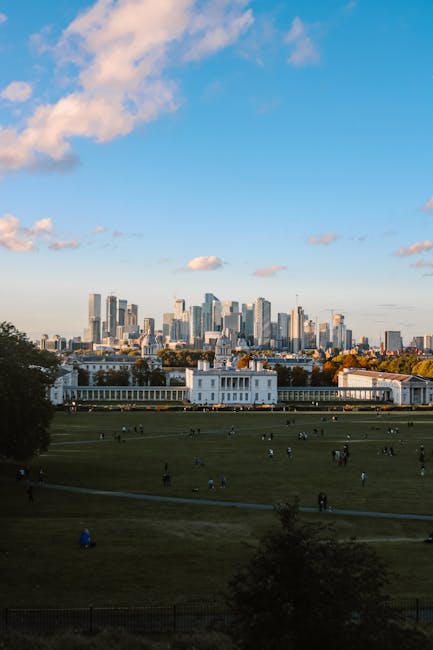 A wide view of Greenwich Park under a partly cloudy sky, featuring lush green grass with numerous visitors relaxing and walking. The central historic building with white columns and a symmetrical faç