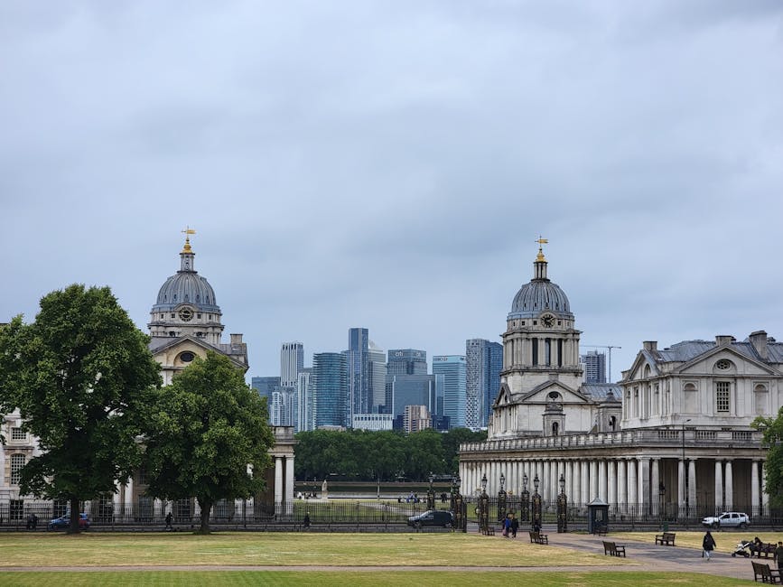 A cityscape view of Greenwich featuring a mix of modern skyscrapers with glass and steel facades, some under construction with cranes visible, set against a clear sky. In the foreground, there is a li